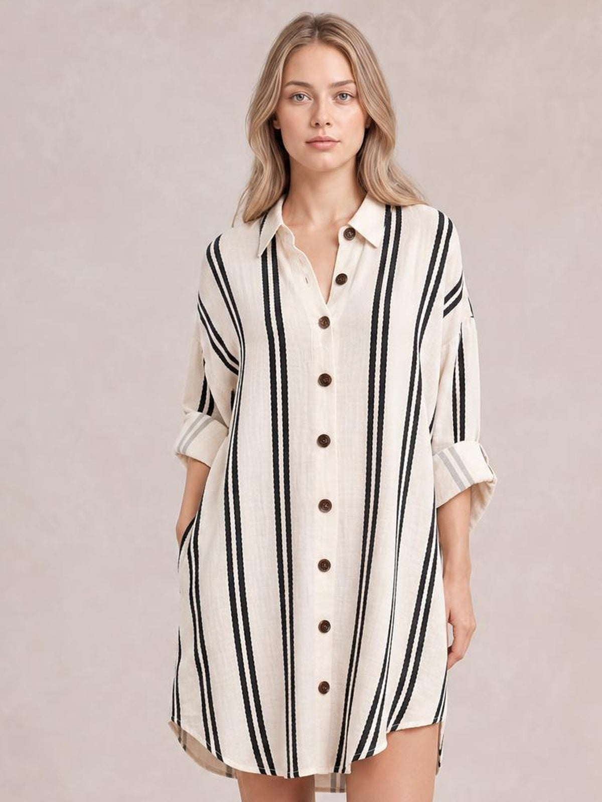Model poses wearing a cream striped button-down shirt dress with vertical black stripes, a collared neckline, front buttons, rolled cuffs, and side pockets.