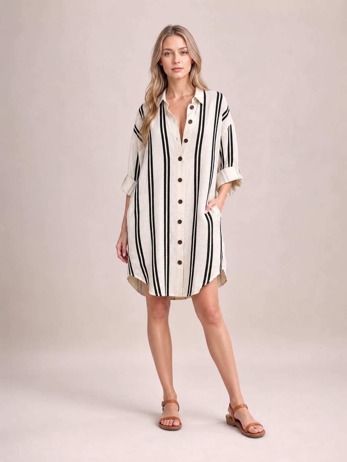 Model wears a cream striped button-down shirt dress with vertical black stripes, collared neckline, rolled sleeves, front buttons, side pockets, and a relaxed short silhouette.