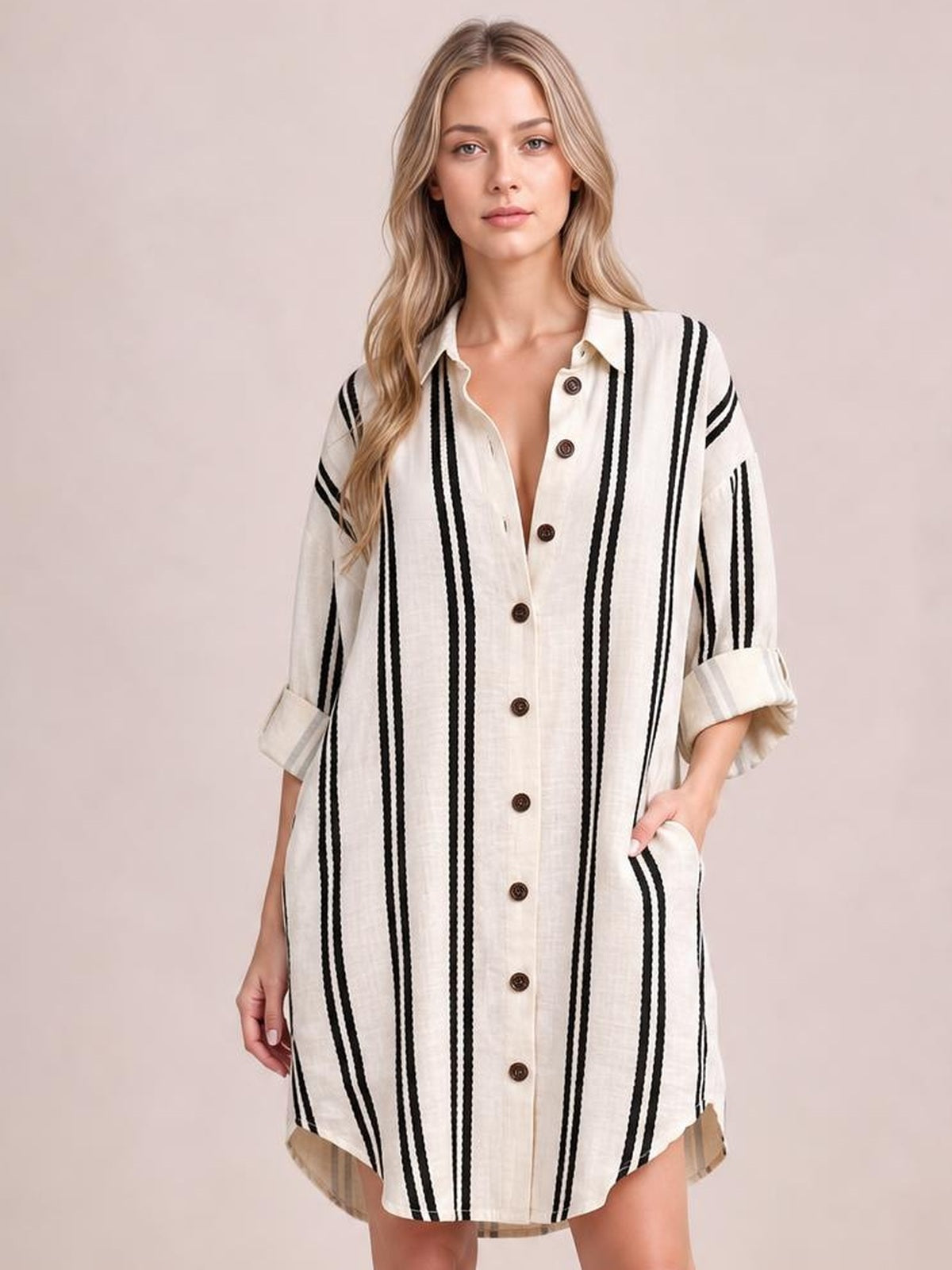 Model poses in a cream striped button-down shirt dress with black vertical stripes, collared neckline, front buttons, rolled sleeves, and side pockets.