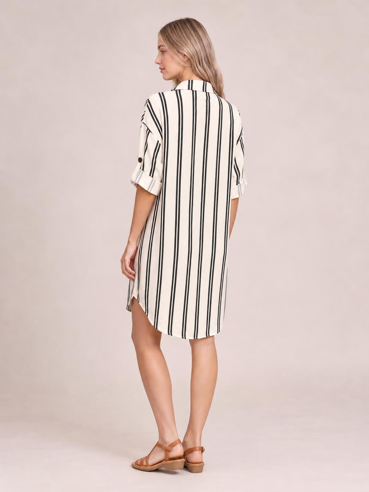 Model shown from the back in a cream shirt dress with black vertical stripes, button-down collar, rolled short sleeves, side slit, and relaxed straight silhouette.
