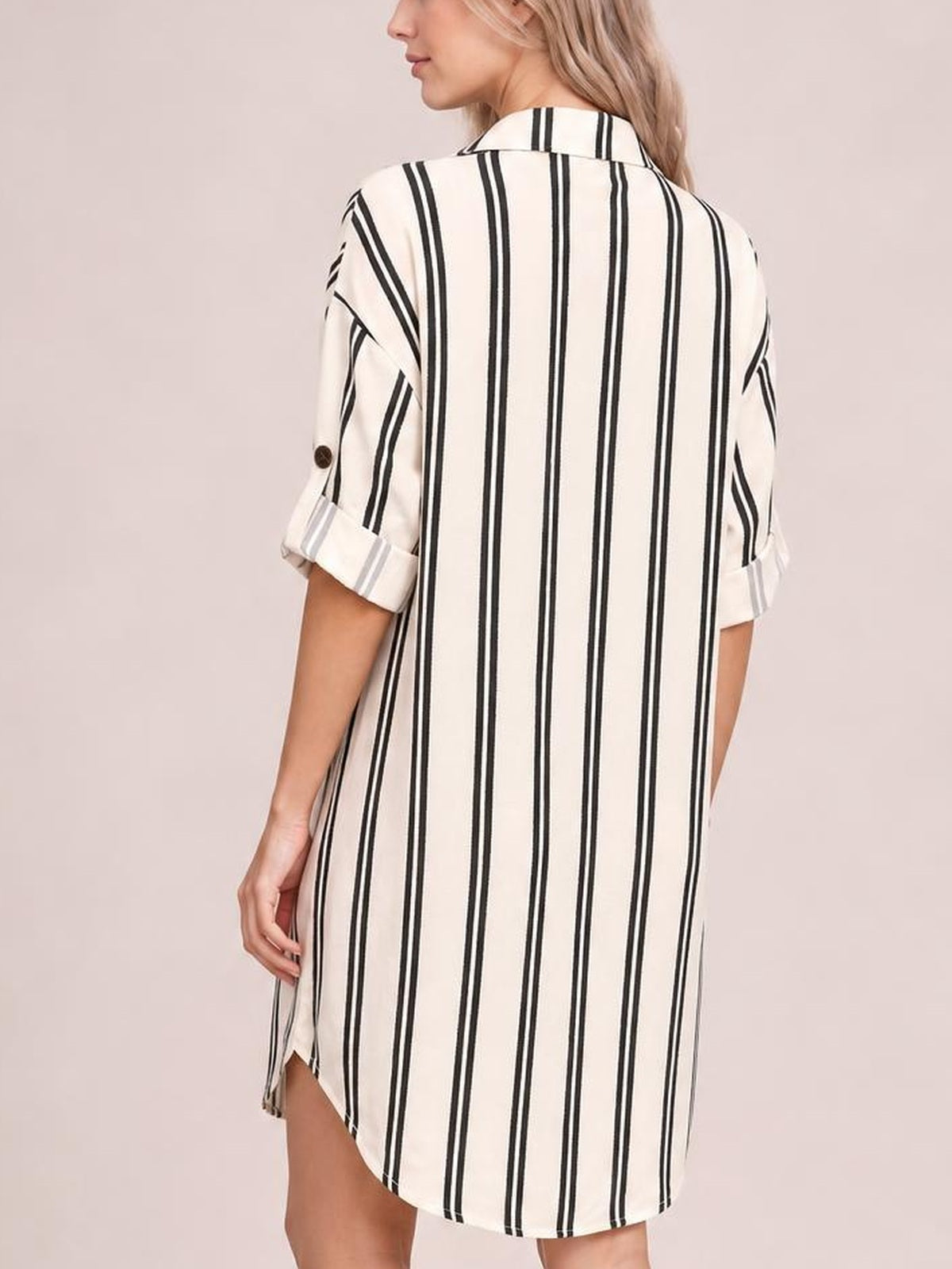 Model turned to show the back of a cream striped button-down shirt dress with black vertical stripes, rolled sleeves, collar, and relaxed straight silhouette.