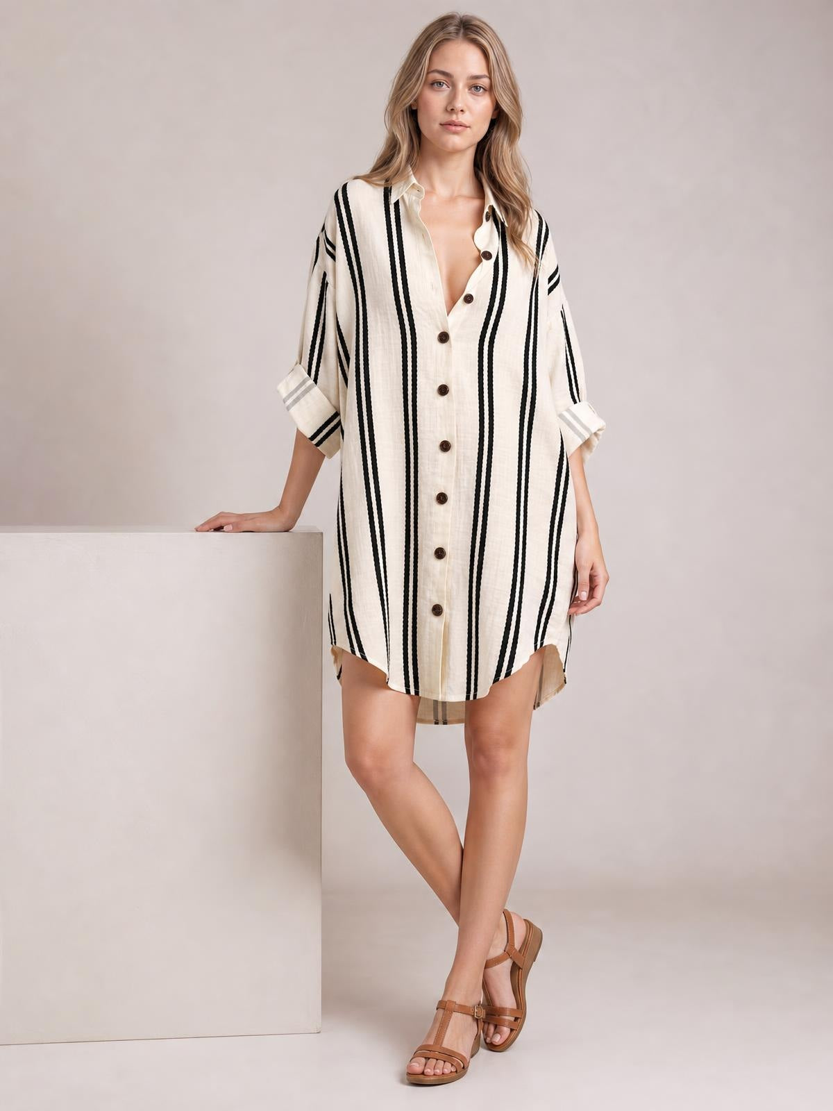A model poses in a relaxed ivory striped button-down shirt dress with black vertical stripes, collar, front buttons, rolled sleeves, and a curved hem.