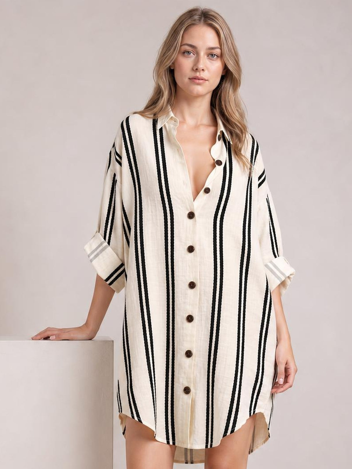 Model wears an oversized cream striped button-down shirt dress with black vertical stripes, rolled sleeves, collared neckline, front buttons, and a curved hem.