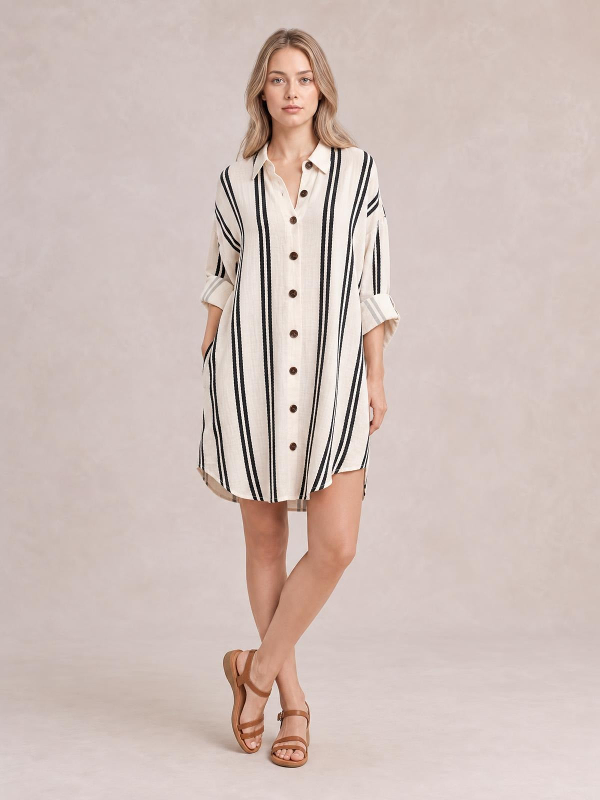 A model poses in a cream-and-black striped button-down shirt dress with a collar, rolled sleeves, front buttons, side pockets, and a relaxed straight silhouette.