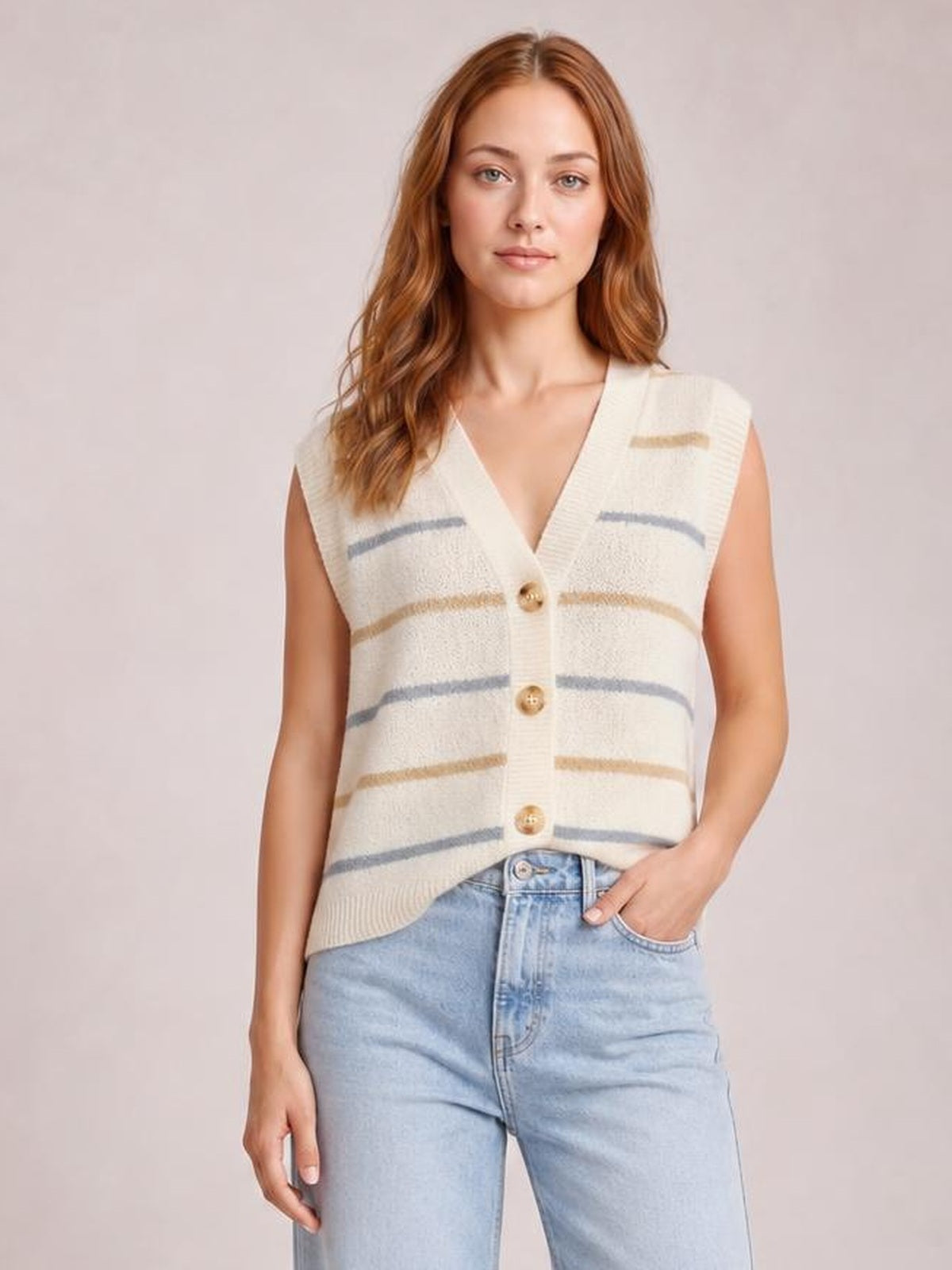 Model wears a cream striped button-down sweater vest with a V-neck, sleeveless cut, relaxed silhouette, and three visible front buttons, posing with one hand in her jeans pocket.