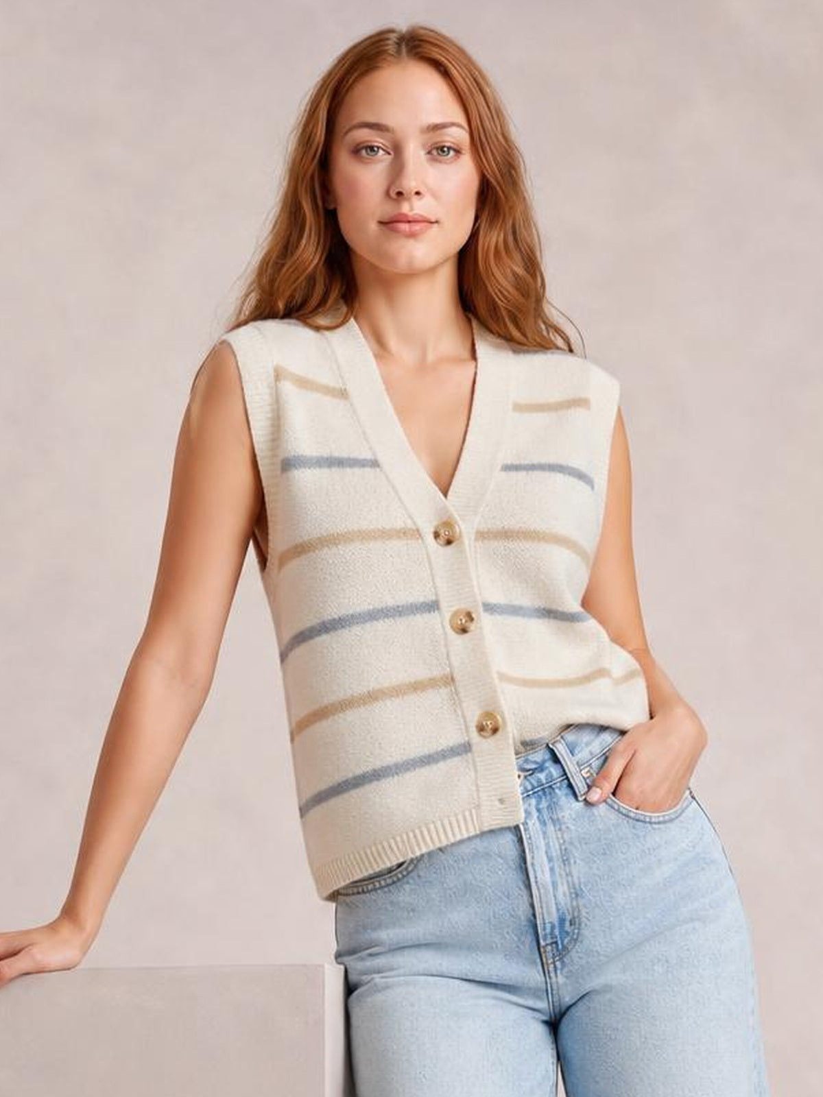 Model wearing a cream striped button-down sweater vest with a V-neck, sleeveless silhouette, ribbed trim, and front buttons, posed with one hand in her jeans pocket.