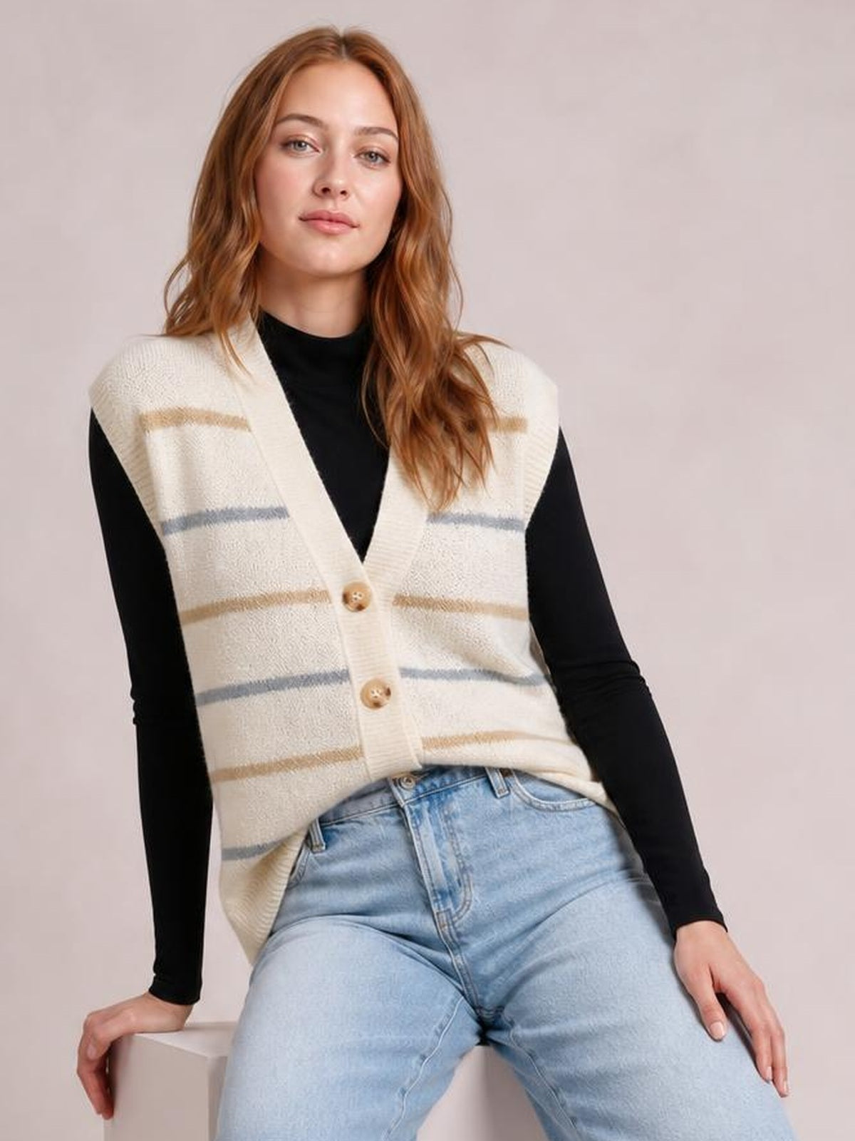 A seated model wears a cream striped button-down sweater vest with a V-neck, sleeveless relaxed silhouette, and beige and gray horizontal stripes over a black top.