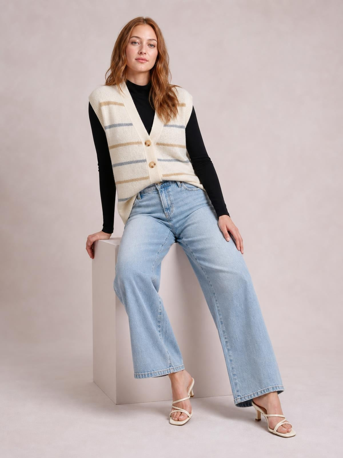 Model seated in a relaxed pose wears a cream striped button-down sweater vest with a V-neck, ribbed trim, and two visible buttons over a black top.