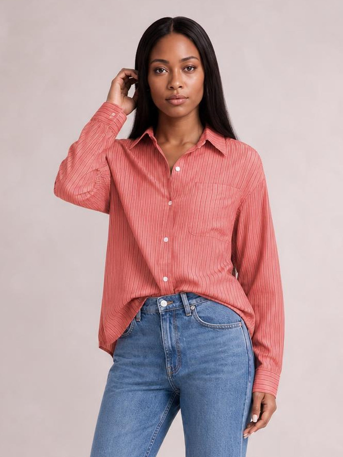 Model wears a coral pink striped button-up long-sleeve shirt with a pointed collar, chest pocket, button front, relaxed silhouette, and one hand raised.