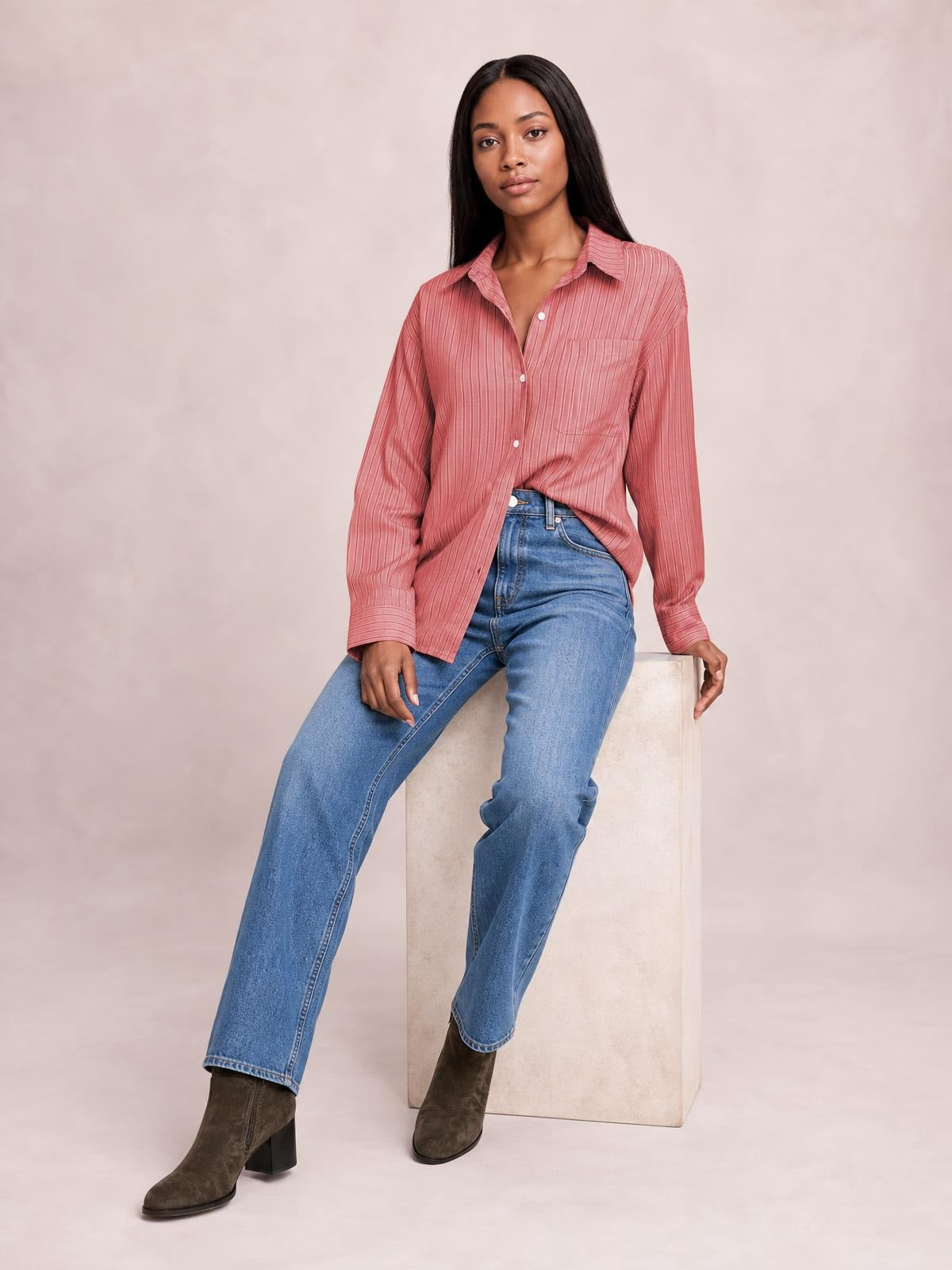 Model seated in a rose pink striped button-up long sleeve shirt with a collared neckline, buttoned chest pocket, relaxed silhouette, and partial tuck.