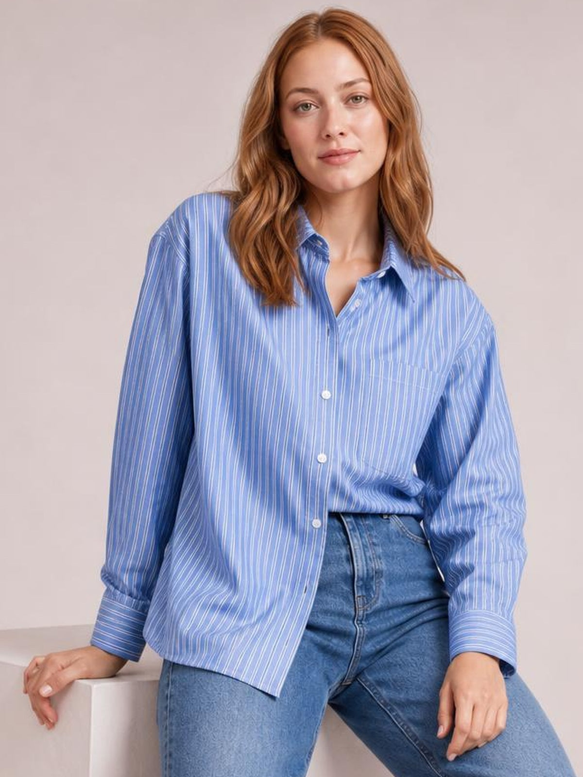Model poses in a blue striped button-up long-sleeve shirt with a collar, button front, buttoned cuffs, relaxed silhouette, and high-waisted jeans.