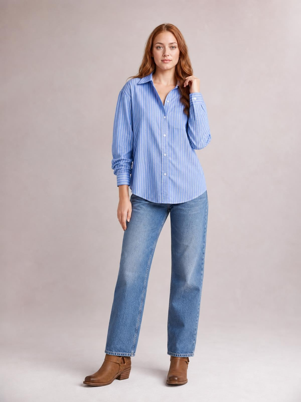 Model wears a blue-and-white striped button-up long-sleeve shirt with a collared neckline, chest pocket, curved hem, and relaxed silhouette while touching her hair.