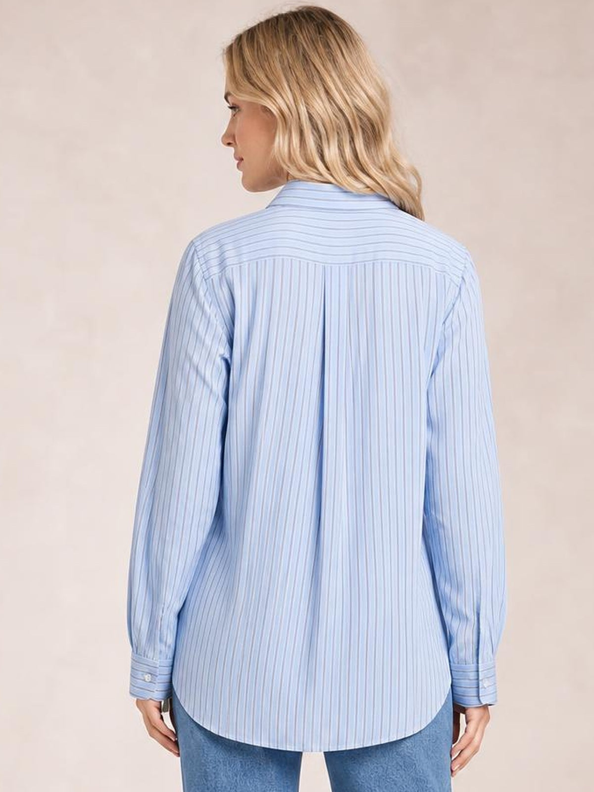 Model shown from the back in a light blue striped button-up long sleeve shirt with a loose silhouette, curved hem, and buttoned cuffs.