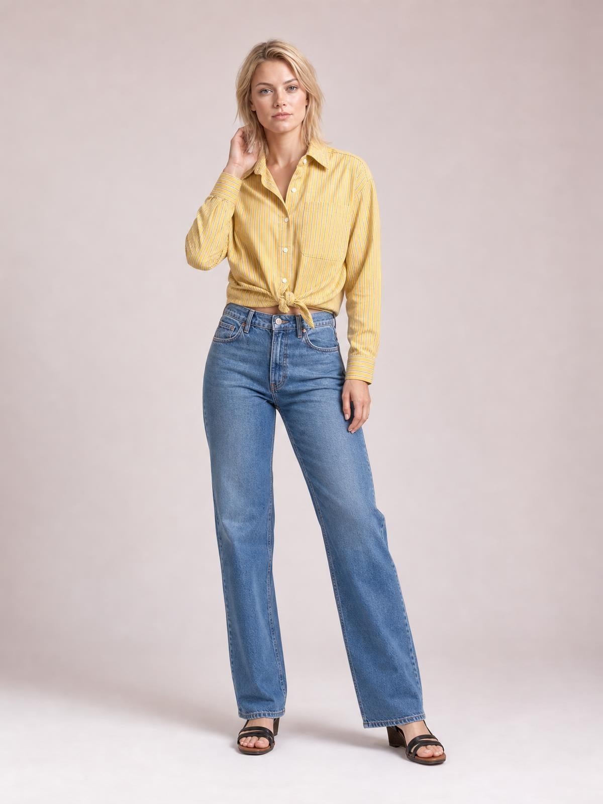 Model wearing a yellow striped button-up long-sleeve shirt with a collared neckline, buttoned chest pocket, and tied waist, posing with one hand at her hair.