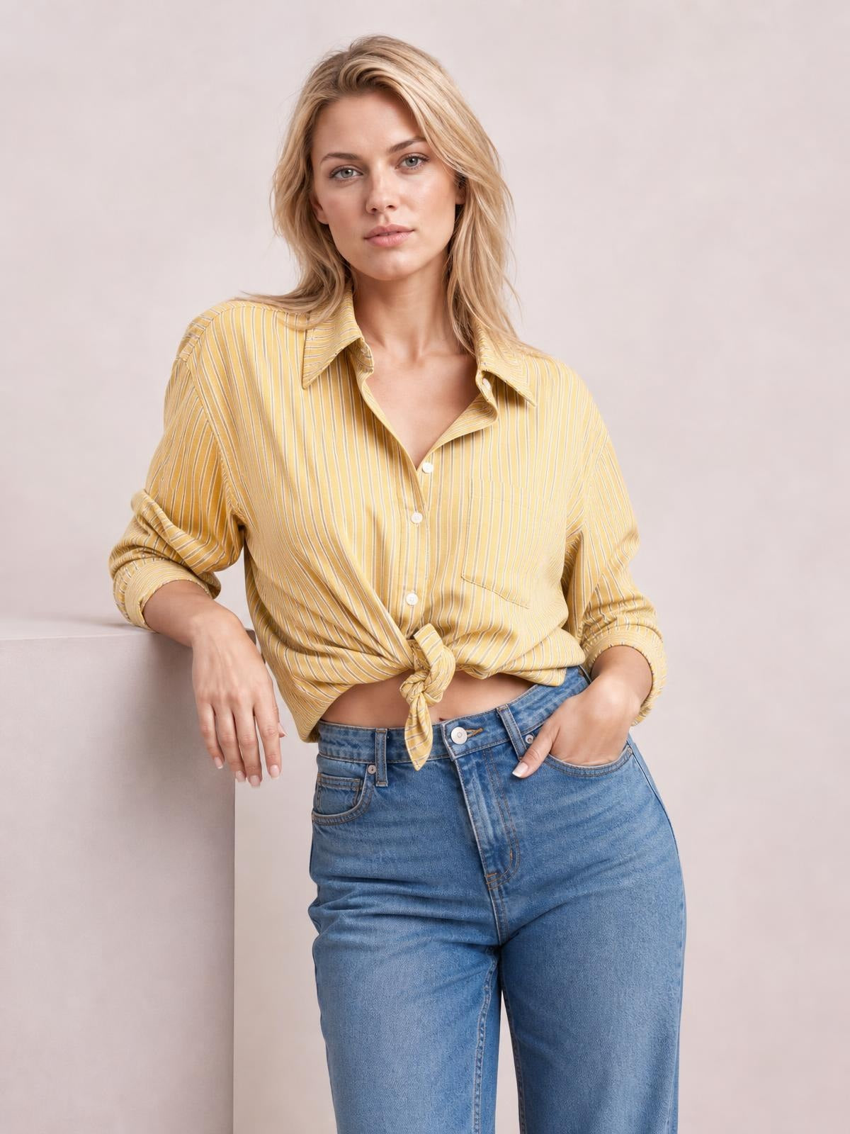 A model leans casually with one hand on a ledge, wearing a mustard yellow striped button-up long sleeve shirt with a collared neckline, button front, rolled cuffs, and tied hem.