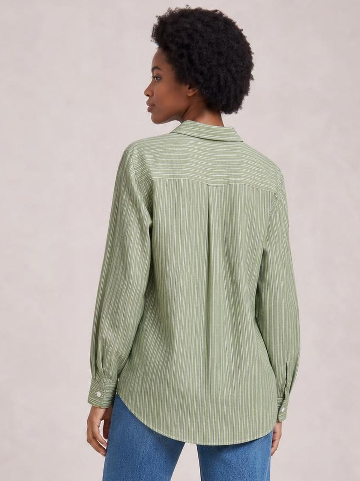 A model stands turned away in a sage green striped button-up long sleeve shirt with a relaxed silhouette, pointed collar, back yoke, and curved hem.
