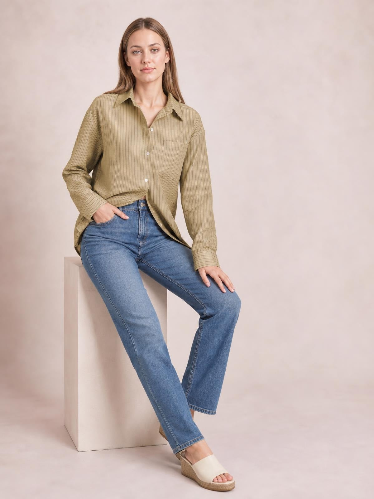 A seated model wears an olive striped button-up long sleeve shirt with a collared neckline, button front, relaxed silhouette, and visible chest pocket.