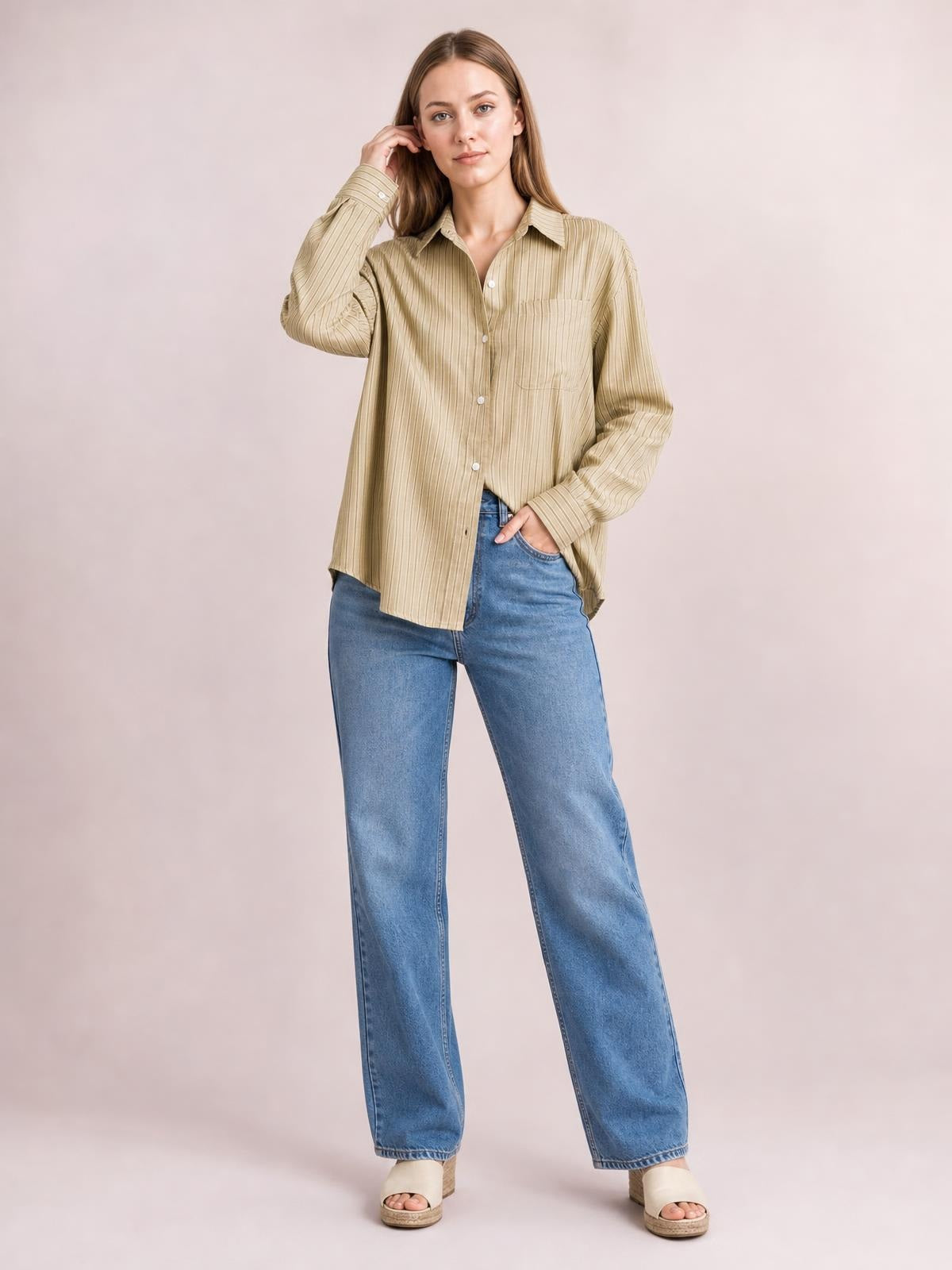 Model wearing a beige striped button-up long-sleeve shirt with a collar, chest pocket, and relaxed silhouette, posing with one hand in her jeans pocket.