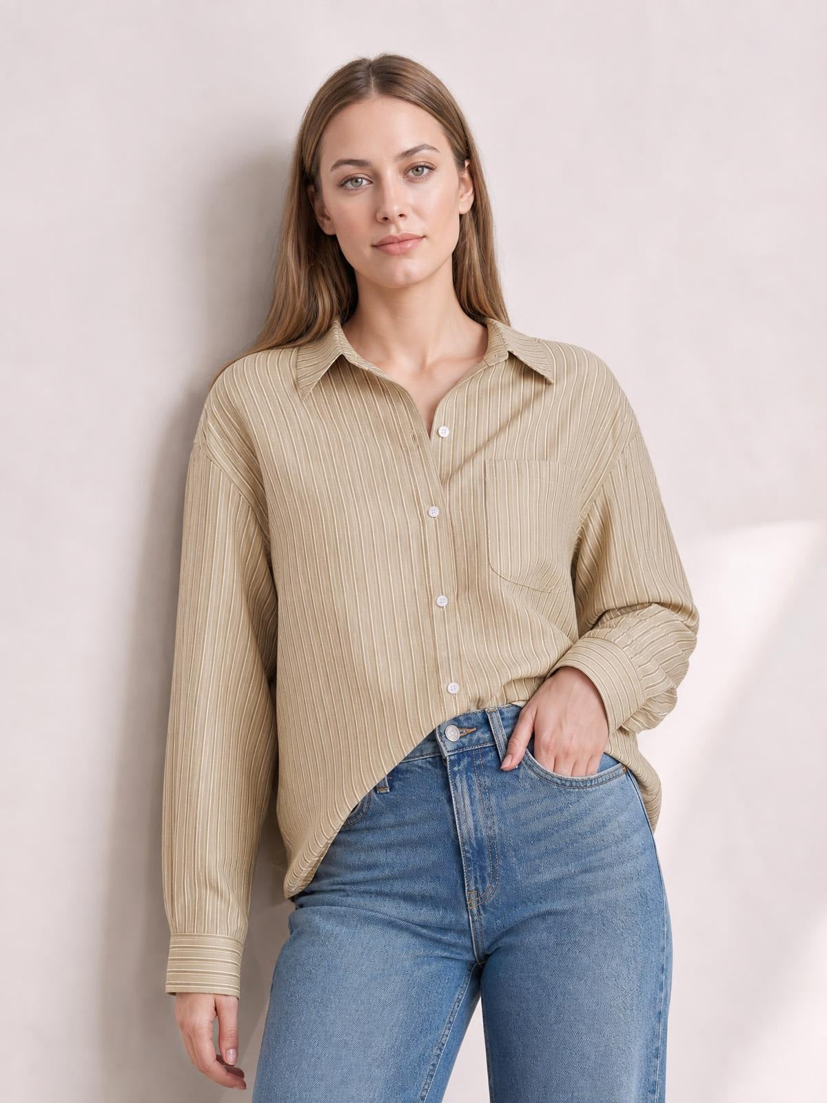 A woman models a beige striped button-up long-sleeve shirt with a collared neckline, front buttons, chest pocket, relaxed silhouette, and one hand in her jeans pocket.