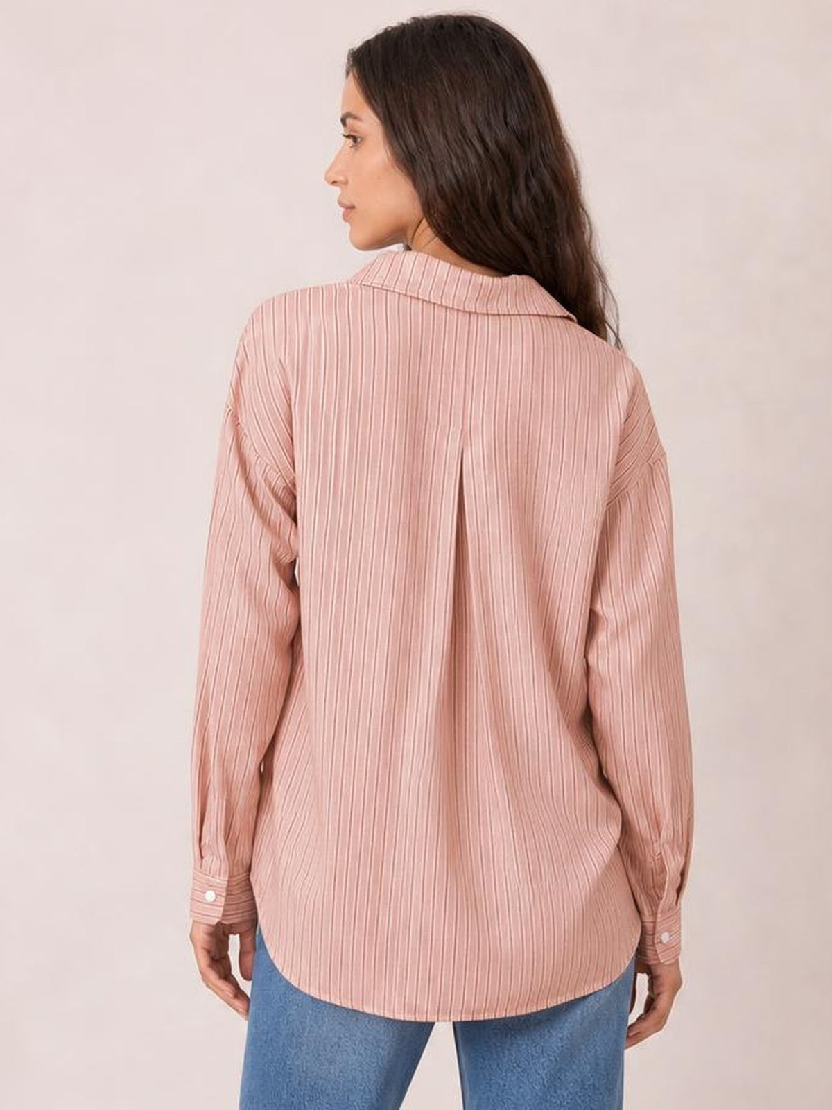 Model shown from the back wearing a pink striped button-up long sleeve shirt with a relaxed silhouette, pointed collar, back pleat, and rounded hem.