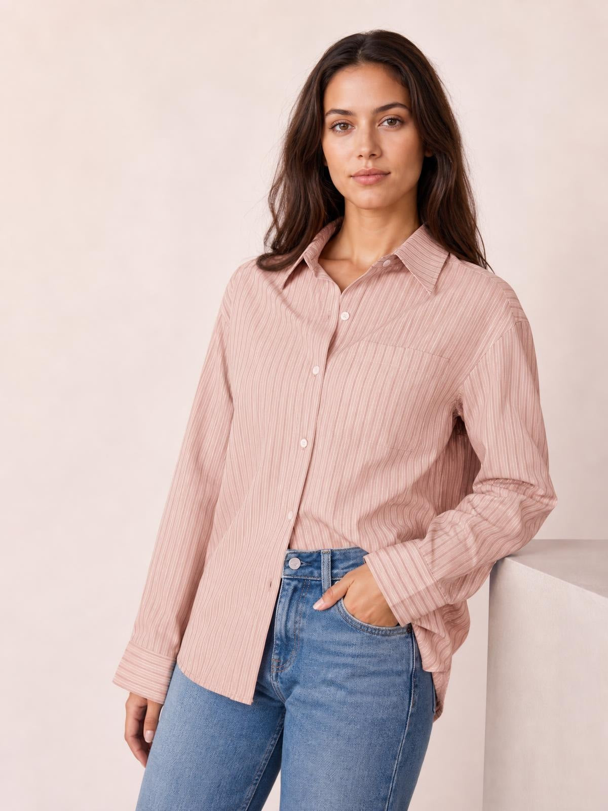 A model stands with one hand in her jeans pocket wearing a blush pink striped button-up long-sleeve shirt with a collar, chest pocket, cuffs, and relaxed silhouette.
