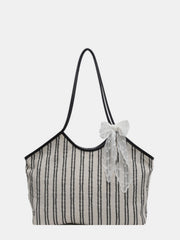 Beige striped canvas tote bag with relaxed vertical black stripes, slim black shoulder straps, a softly structured silhouette, and a feminine lace bow detail.