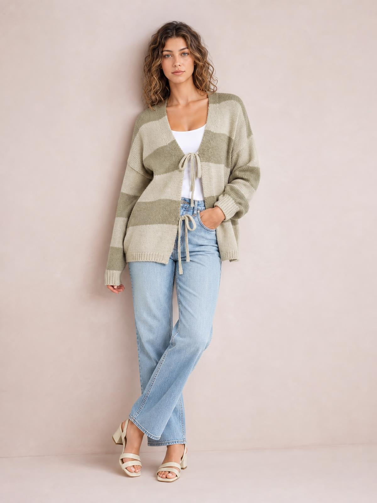 A model stands cross-legged in a relaxed striped color-block sweater cardigan with olive green and cream bands, long sleeves, and a tied V-neck front.