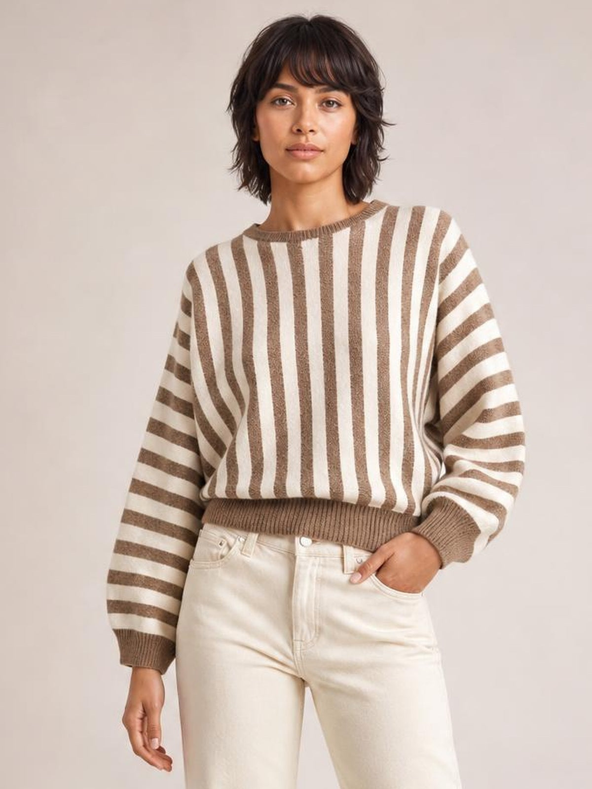Model wearing a brown and cream striped crew neck sweater with a relaxed silhouette, ribbed collar, cuffs, and hem, posing with one hand in her pocket.