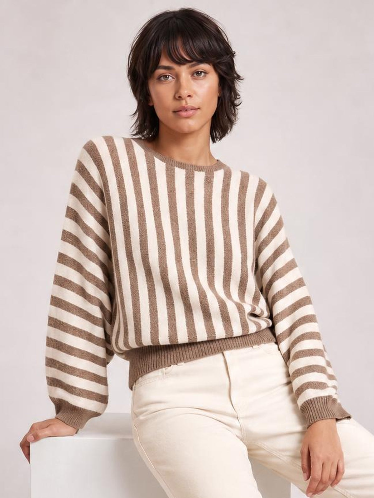 Model seated in a relaxed pose wearing a brown and cream striped crew neck sweater with a loose silhouette, ribbed cuffs, neckline, and hem.