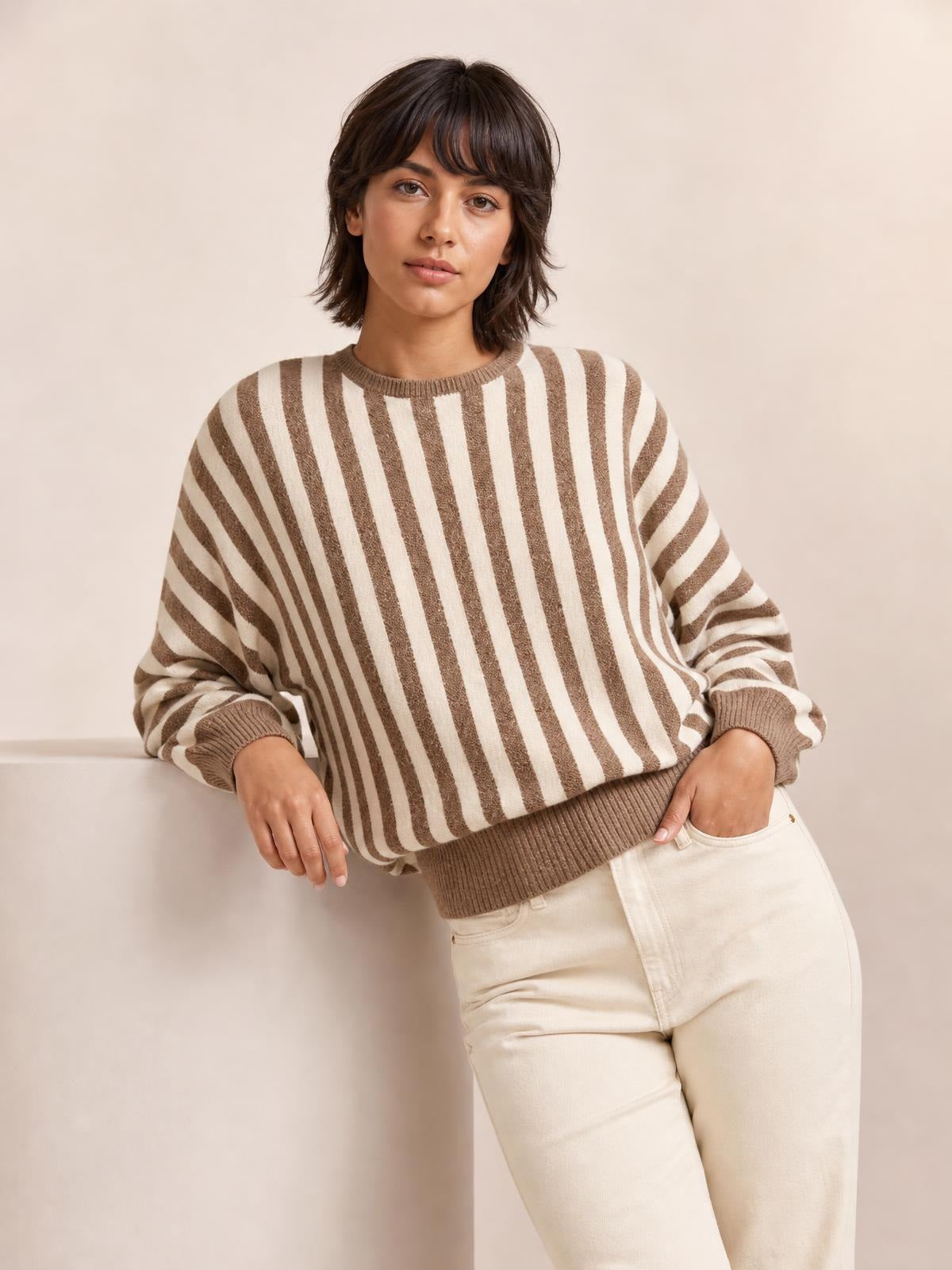 Model wearing a brown and cream striped crew neck sweater with a relaxed silhouette, ribbed neckline, cuffs, and hem, posing with one hand in her pocket.