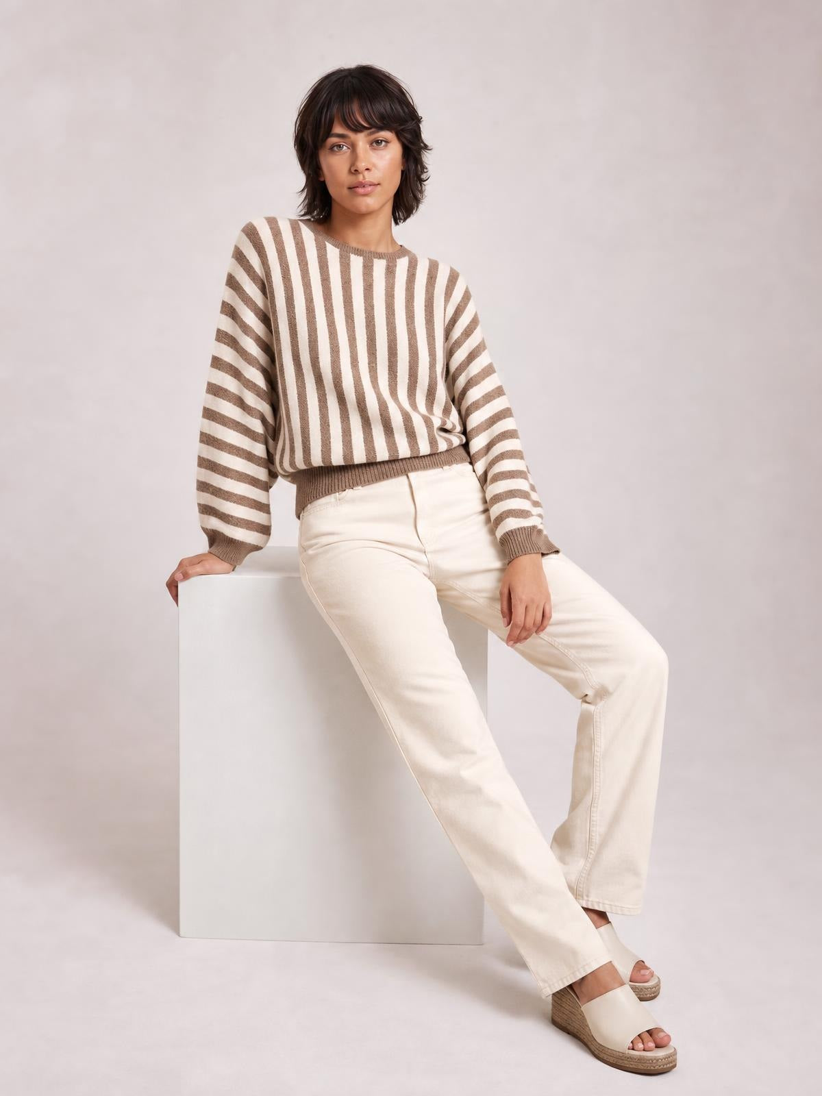 Model seated in a relaxed pose wearing a brown and cream striped crew neck sweater with ribbed trim, long sleeves, and a slightly loose silhouette.