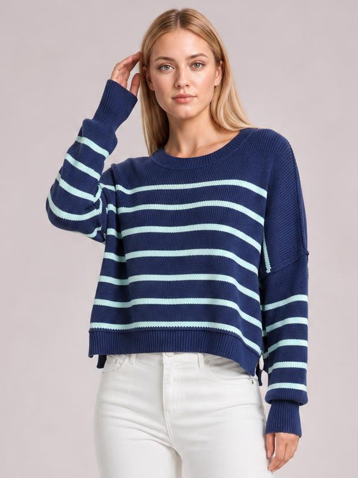 Model poses with one hand near her hair wearing a navy striped dropped-shoulder round-neck pullover sweater with long lantern sleeves and a relaxed silhouette.