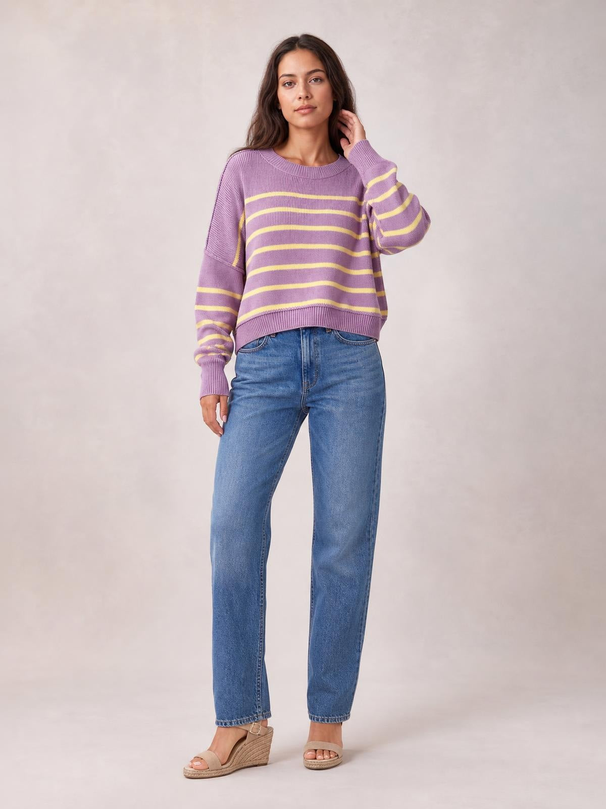 Model wears a lilac and yellow striped round-neck pullover sweater with dropped shoulders, lantern sleeves, ribbed cuffs and hem, posing with one hand near her hair.