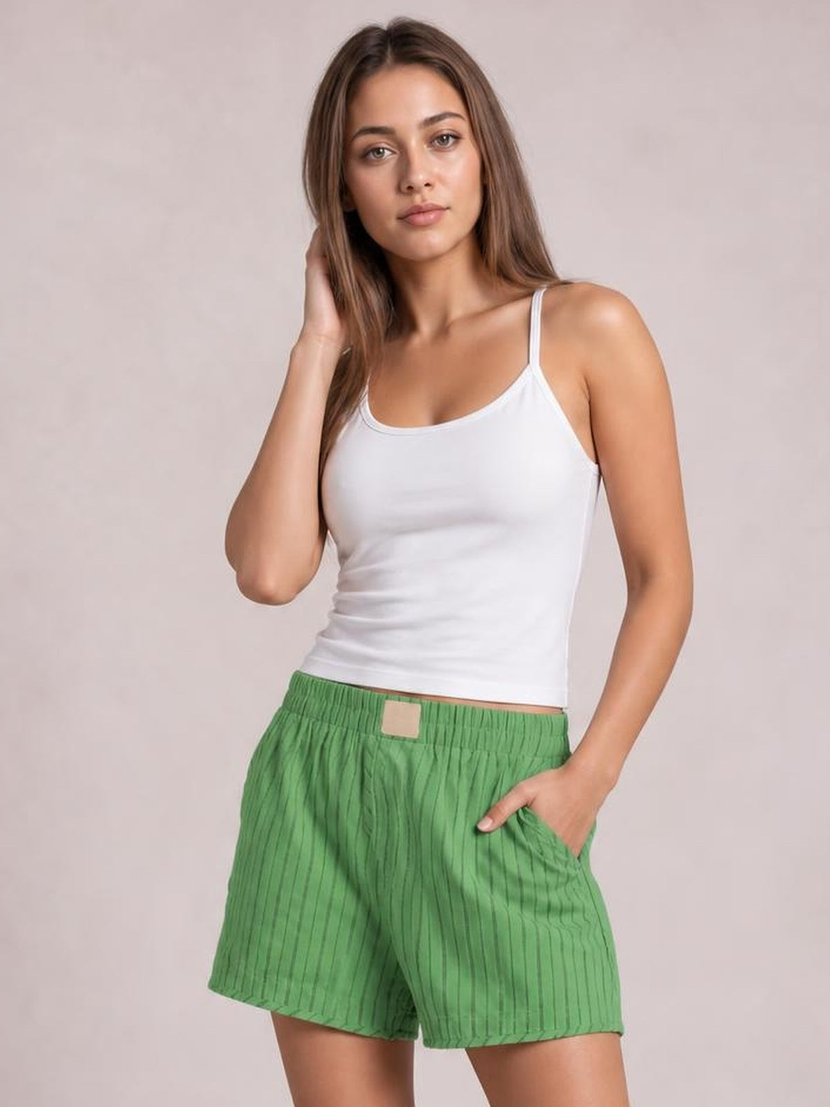 A woman modeling a white ribbed camisole tank top and green pinstriped shorts against a light beige studio background.