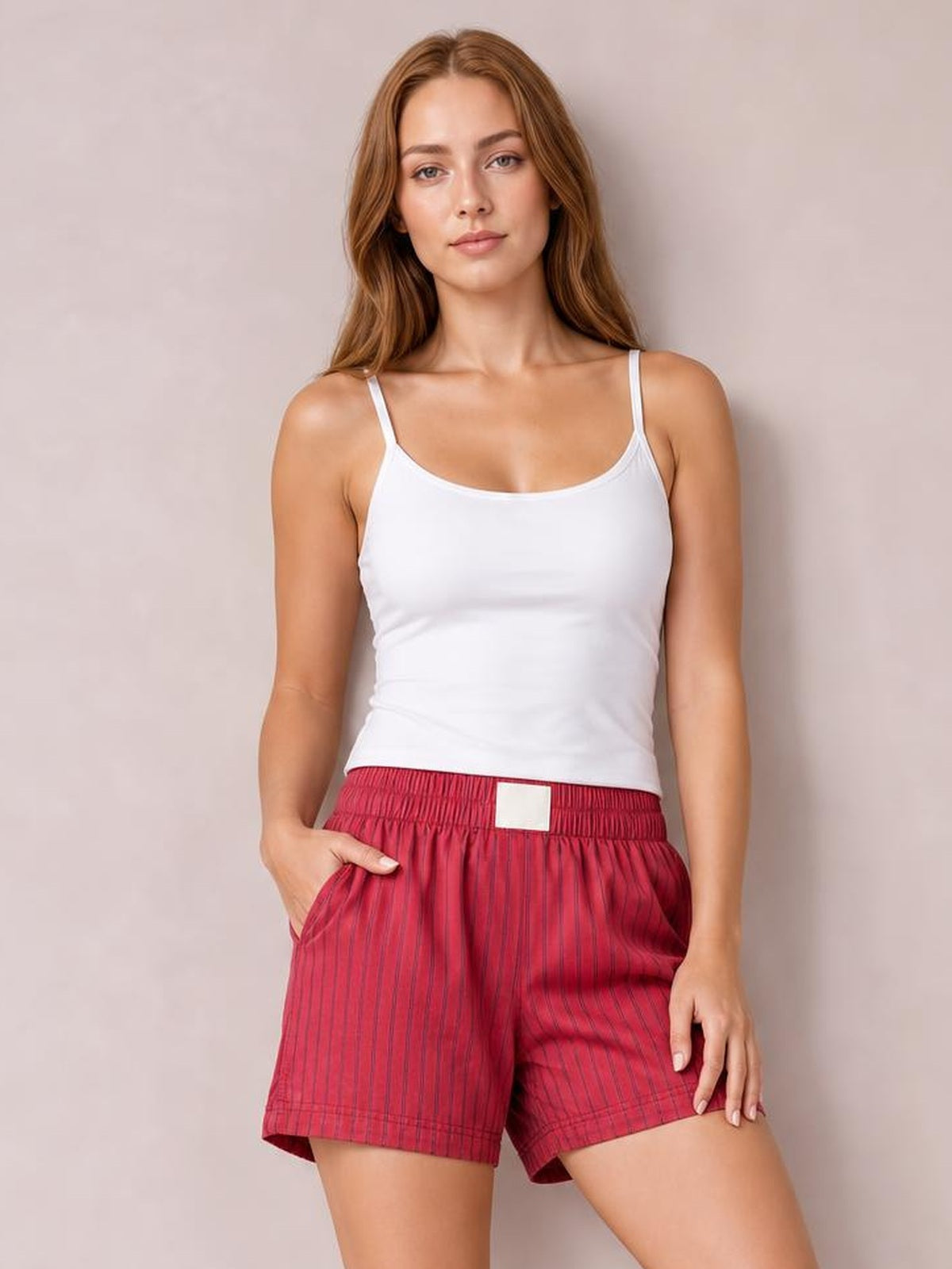 A woman modeling a white spaghetti-strap tank top and red pinstriped shorts against a neutral beige studio backdrop.