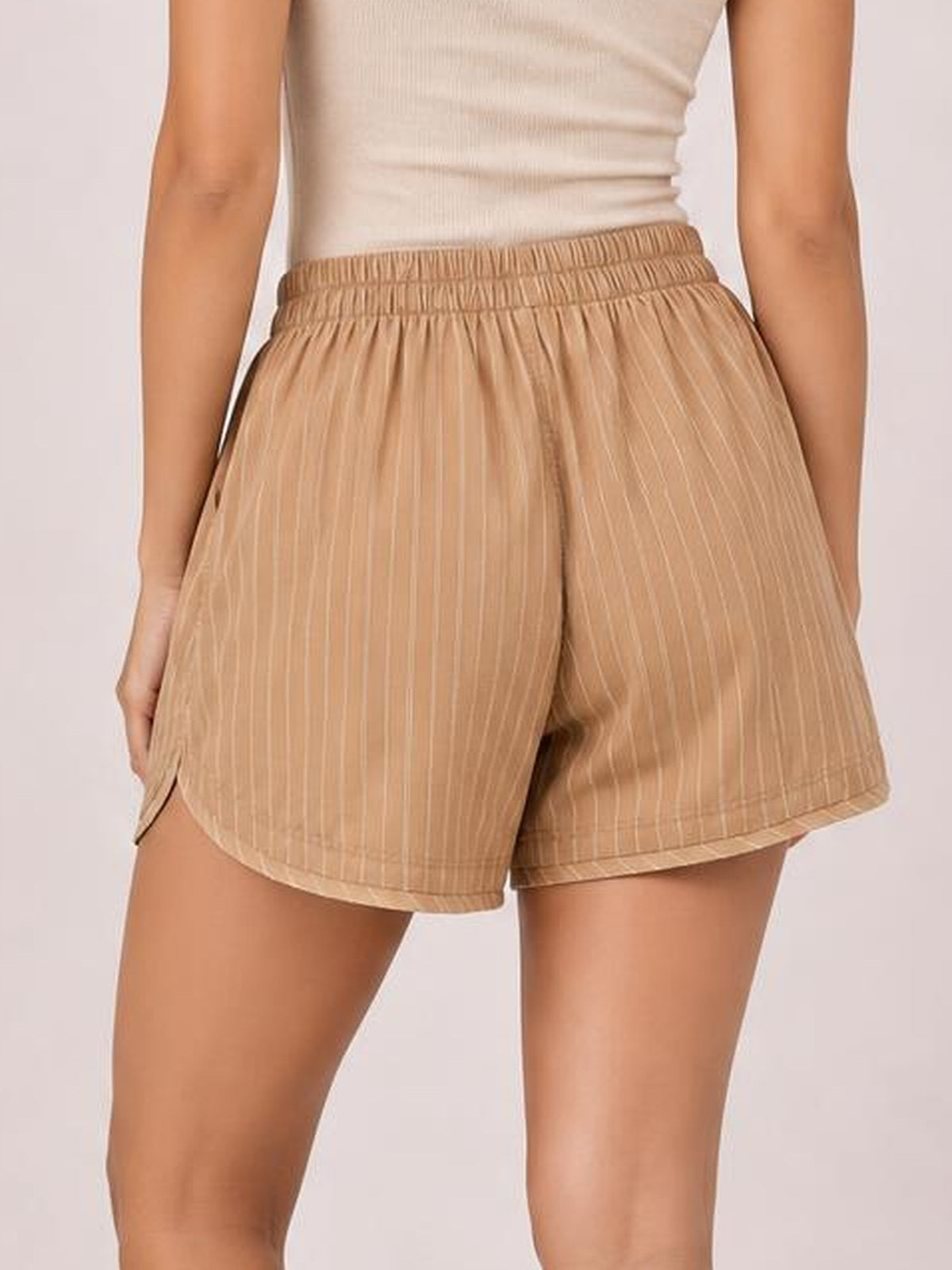 Model wearing tan striped elastic-waist shorts with a relaxed silhouette, side pockets, and curved hems, shown from the back.