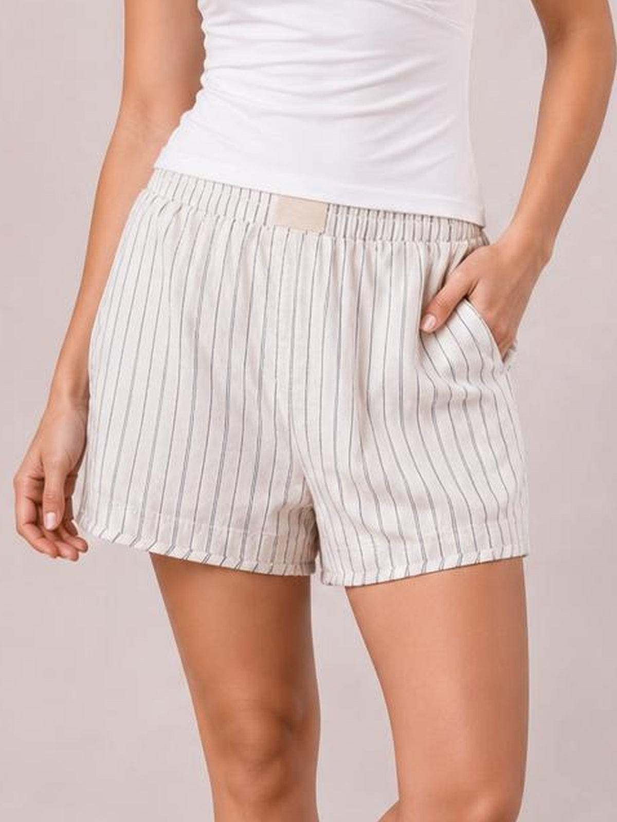 Model wears white striped elastic-waist shorts with a relaxed silhouette, vertical pinstripes, and side pockets, posing with one hand tucked into a pocket.