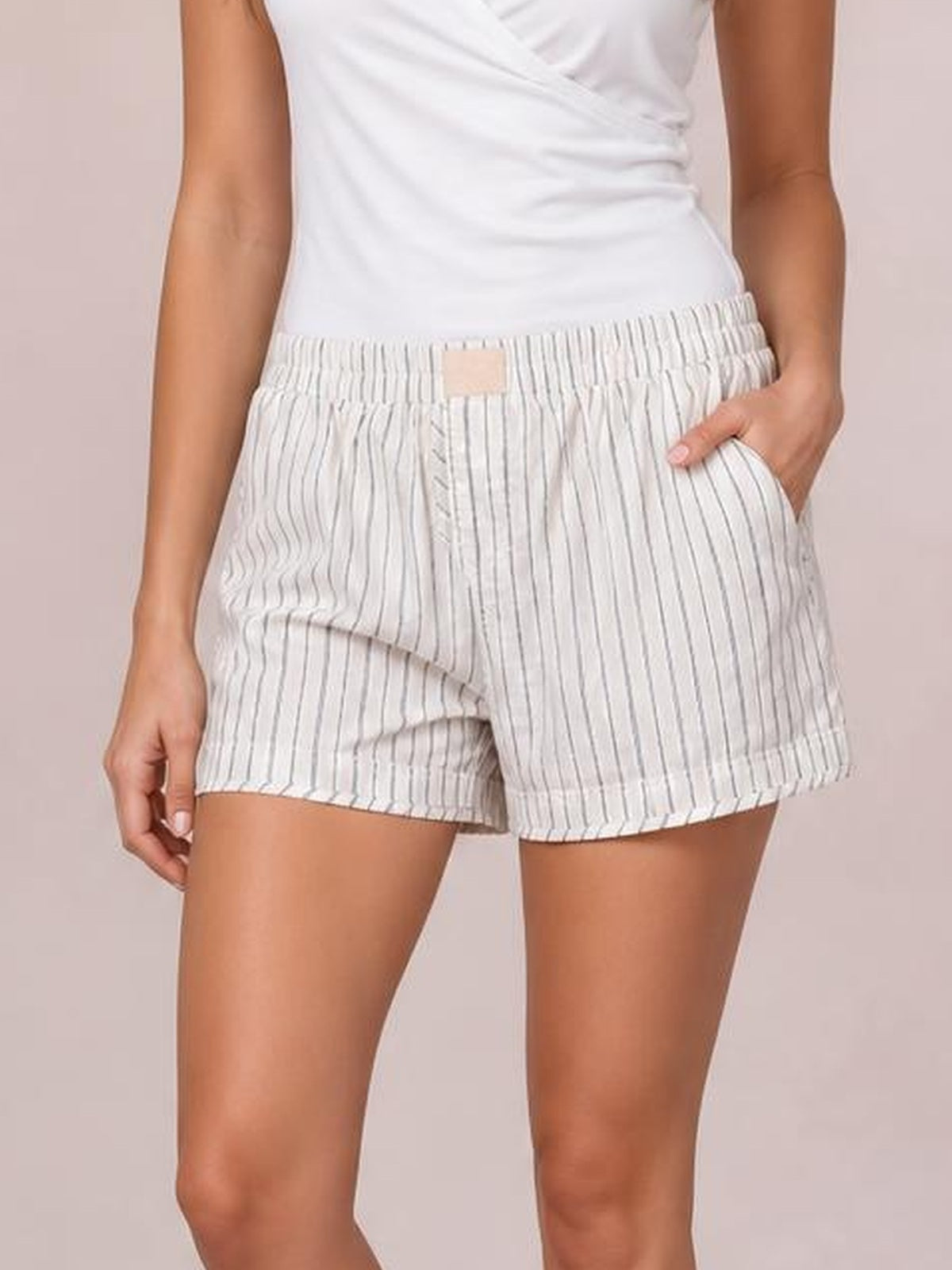 Model wears white striped elastic waist shorts with a relaxed silhouette, visible side pockets, and a hand resting in one pocket.