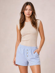 A woman modeling a beige sleeveless knit top and light blue and white striped shorts with gold buttons, set against a solid beige studio background.
