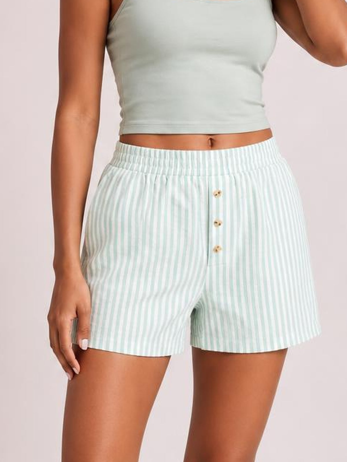 Model wears opaque light mint-and-white striped shorts with an elastic waist, relaxed silhouette, and decorative front buttons, standing with one hand at her side.