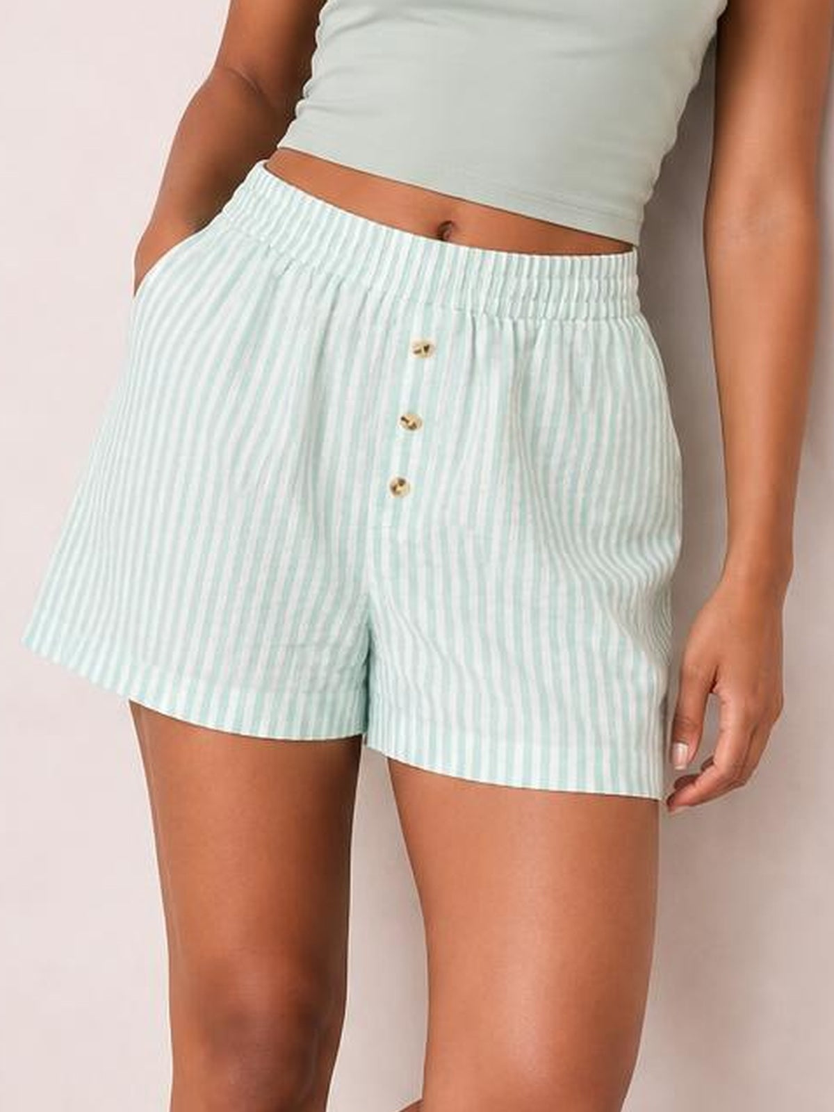 Model wearing opaque light mint-and-white striped elastic waist shorts with a relaxed silhouette, straight hems, and decorative front buttons, standing with one hand in a pocket.