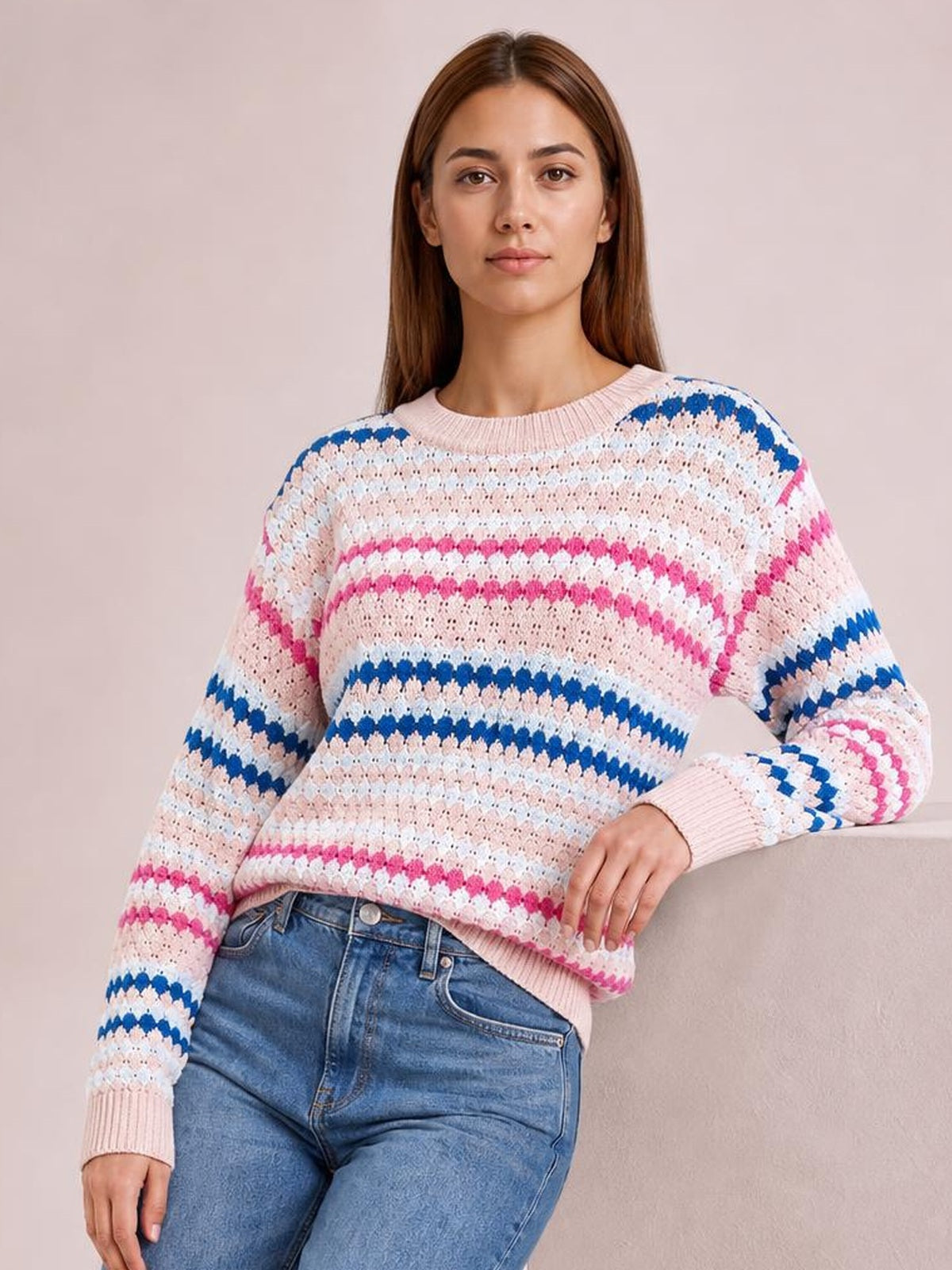 Model wearing a relaxed striped geometric pattern sweater in pink, white, and blue with ribbed crew neckline, cuffs, and hem, posing with one hand on her hip.