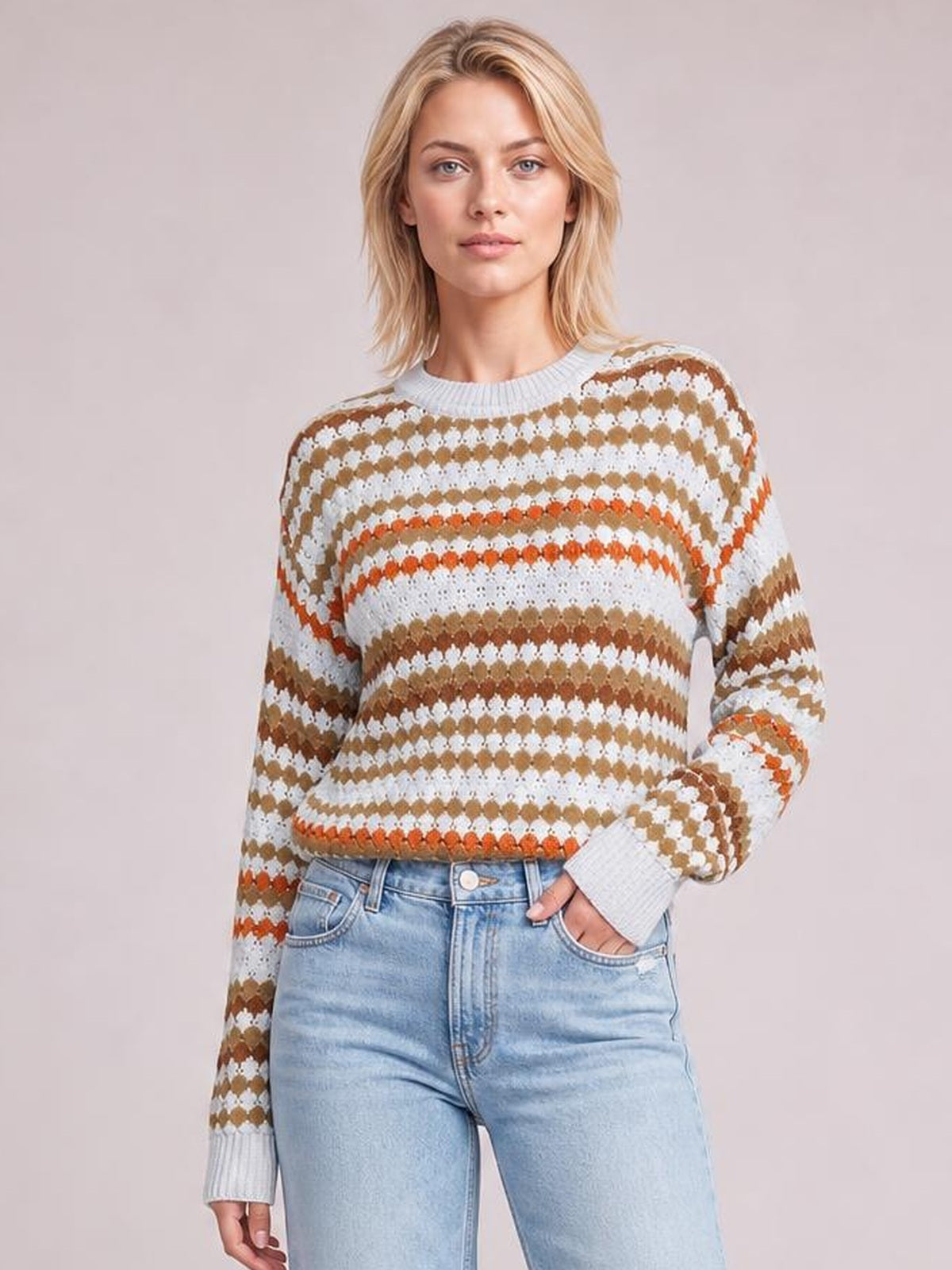 Woman wearing a relaxed striped geometric pattern sweater in white, tan, and orange with a ribbed crew neck and cuffs, one hand in her jeans pocket.