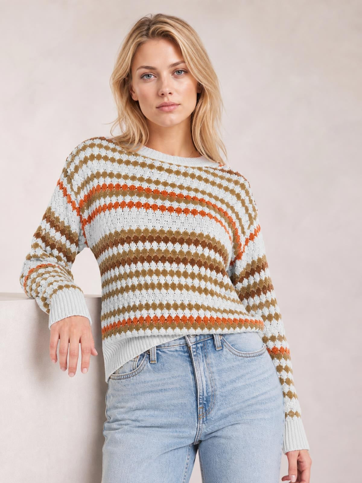 Model wearing a light gray striped geometric pattern sweater with brown and orange accents, ribbed crew neck, cuffs and hem, relaxed silhouette, leaning with one hand resting.