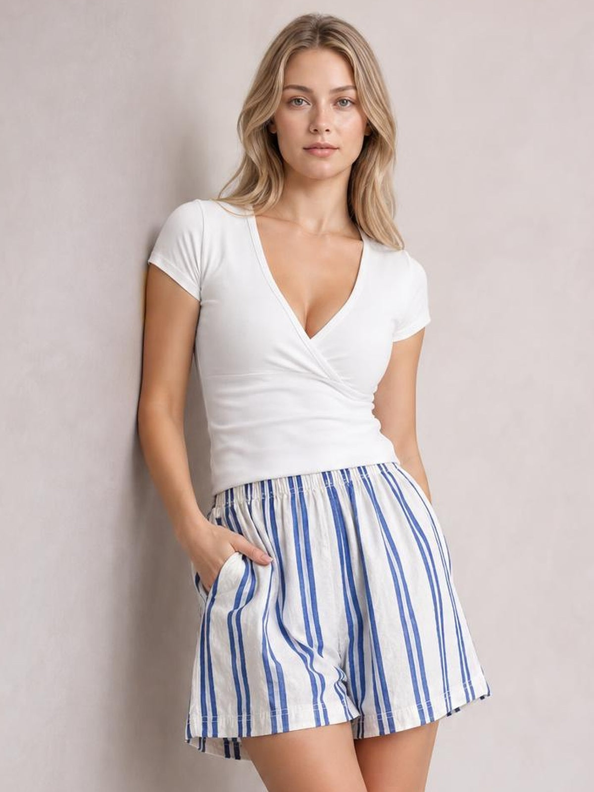 A woman modeling a white short-sleeve wrap-style top and blue-and-white vertically striped shorts against a plain light beige wall.