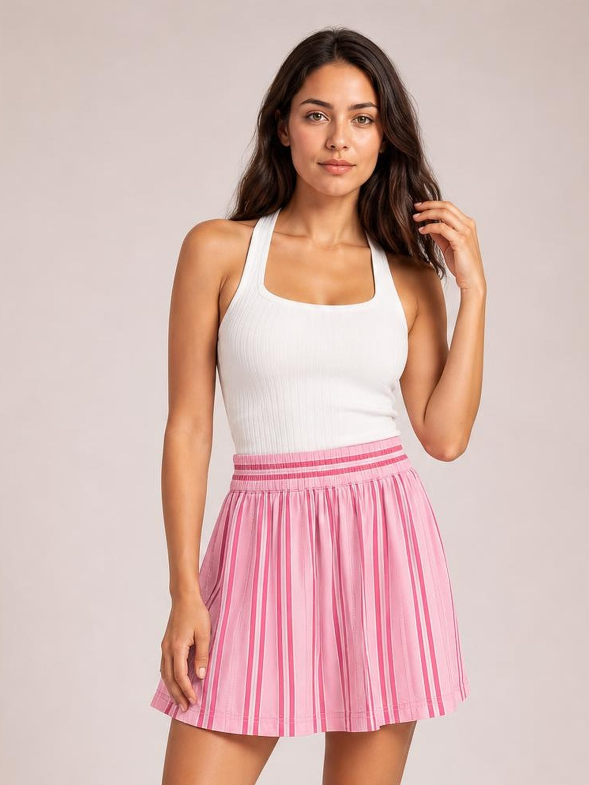 A woman modeling a white ribbed tank top and a pink pleated tennis skirt with vertical pink stripes, set against a solid light beige studio background.