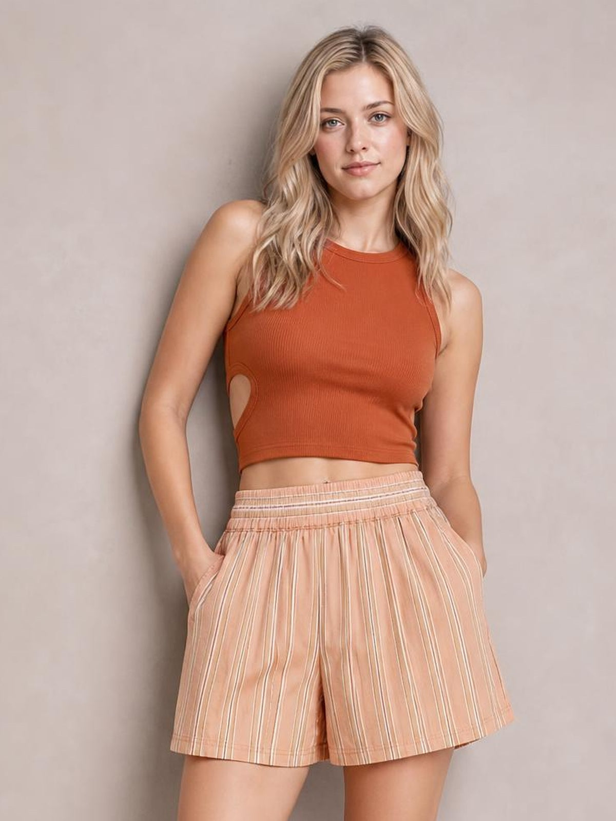 A woman modeling a sleeveless rust-orange crop top and peach striped shorts against a plain beige studio background.