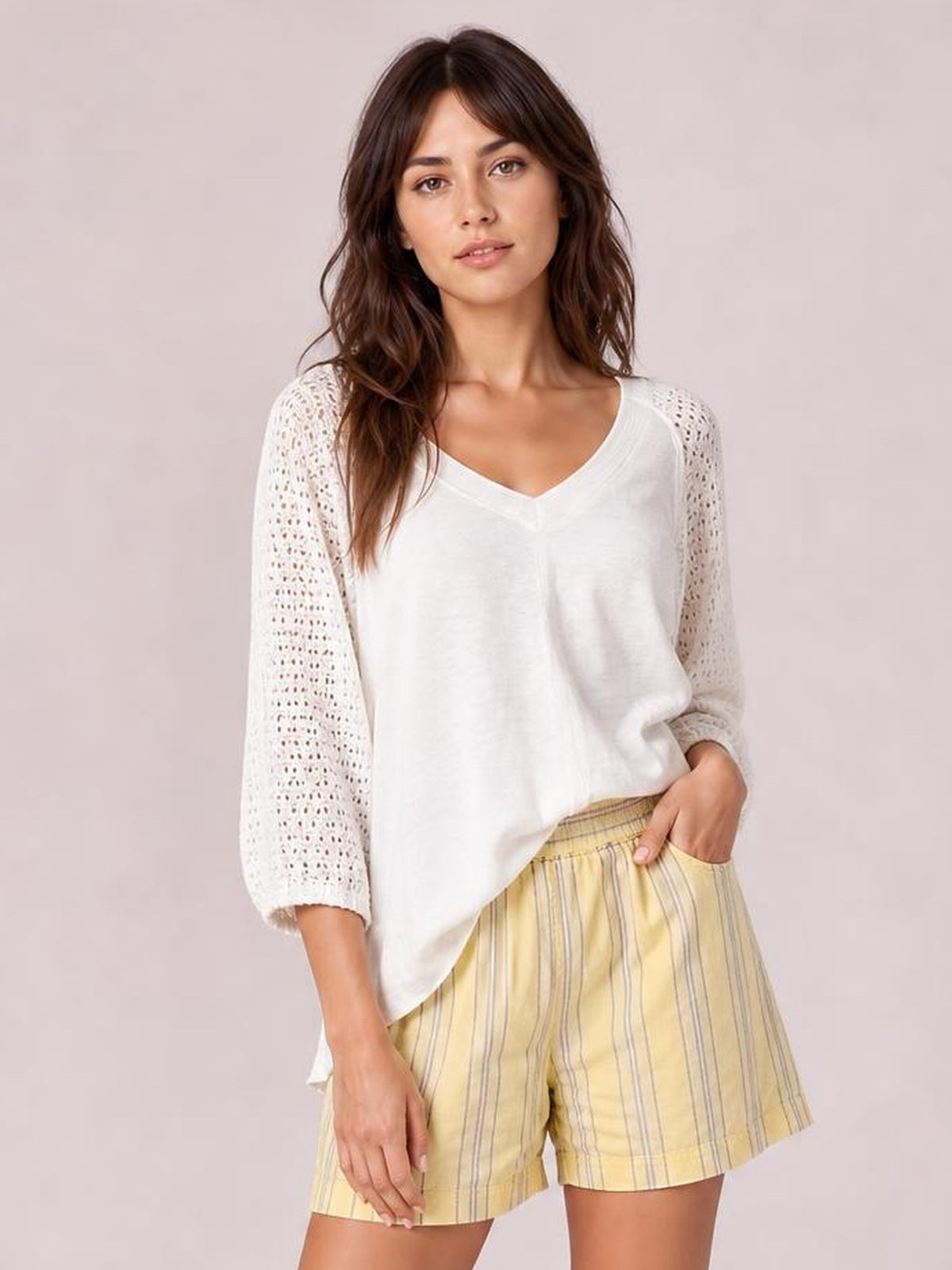 A woman modeling a white knit top with textured, openwork sleeves and yellow-and-white striped shorts against a solid pale pink background.