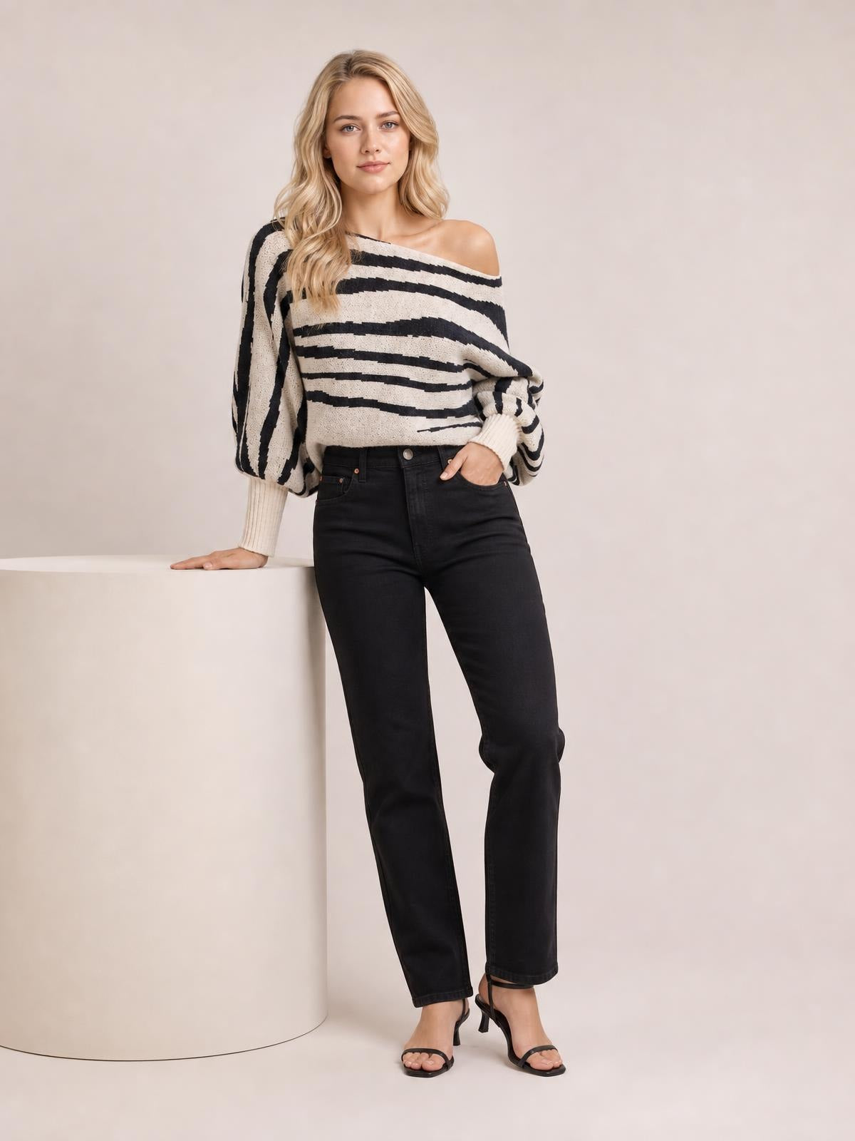 Model wears a cream and black striped sweater with a relaxed off-shoulder neckline, balloon lantern sleeves, and ribbed cuffs while leaning on a pedestal.