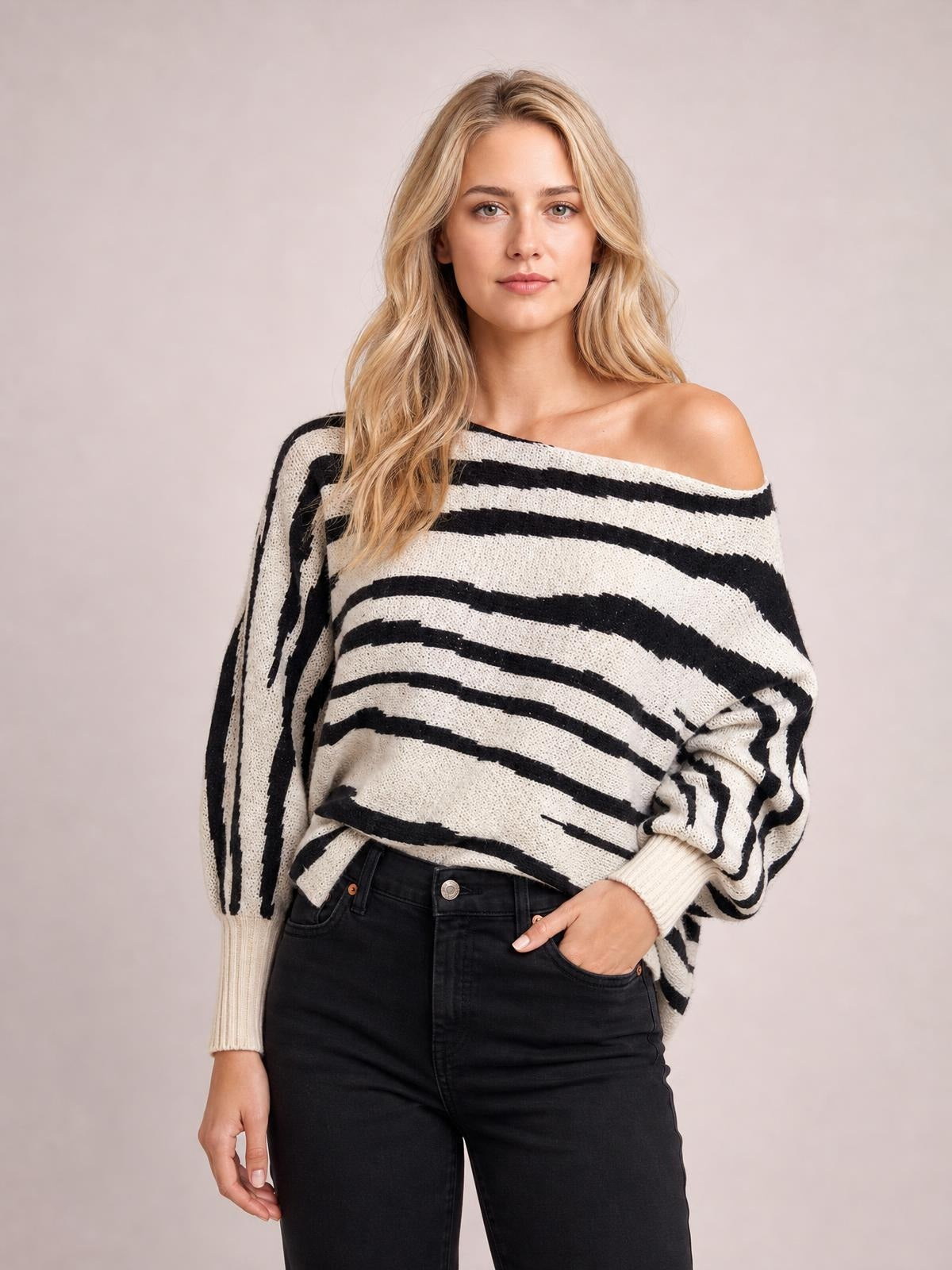 A model wears a cream and black striped sweater with an off-shoulder neckline, relaxed silhouette, lantern sleeves, and ribbed cuffs while posing with one hand in her pocket.
